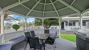 A covered outdoor patio with a table and chairs, a grill, and a view of a residential street. - Phillip Island Cottages (Cowes)