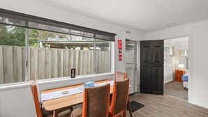 A room with a dining table and chairs, a window with a view of a wooden fence, and a door leading to another room. - Phillip Island Cottages (Cowes)