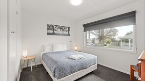 A bedroom with a bed, a bedside table, a lamp, a window with a view, and a painting on the wall. - Phillip Island Cottage 1 (Cowes)