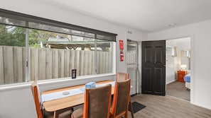 A room with a dining table and chairs, a window with a view of a wooden fence, and a door leading to another room. - Phillip Island Cottage 4 (Cowes)