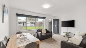 A modern living room with a dining area, a sofa, a TV, and a view of the outside. - Phillip Island Cottage 5 (Cowes)