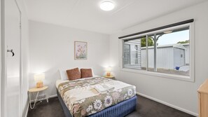 A bedroom with a bed, two bedside tables, a window, and a door. - Phillip Island Cottage 5 (Cowes)