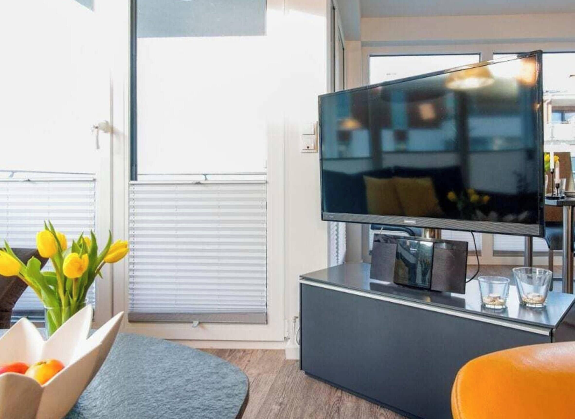 A modern living room with a flat-screen TV on a black stand, a glass coffee table, a blue rug, and a flower vase with yellow flowers.