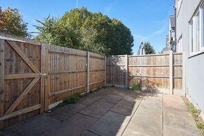 A paved patio area with a wooden fence and a white building in the background. - 3BR Flat in Bromley |Near Local Stores & Resto | ST (London)
