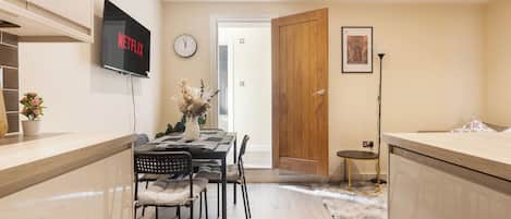 A modern kitchen with a flat-screen TV, a dining area with a wooden table and chairs, and a hallway leading to a door.