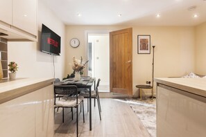 A modern kitchen with a flat-screen TV, a dining area with a wooden table and chairs, and a hallway leading to a door. - 3BR Flat in Bromley |Near Local Stores & Resto | ST (London)