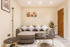 A living room with a sofa, ottoman, two small tables, a floor lamp, a painting, a door, and a potted plant. - 3BR Flat in Bromley |Near Local Stores & Resto | ST (London)