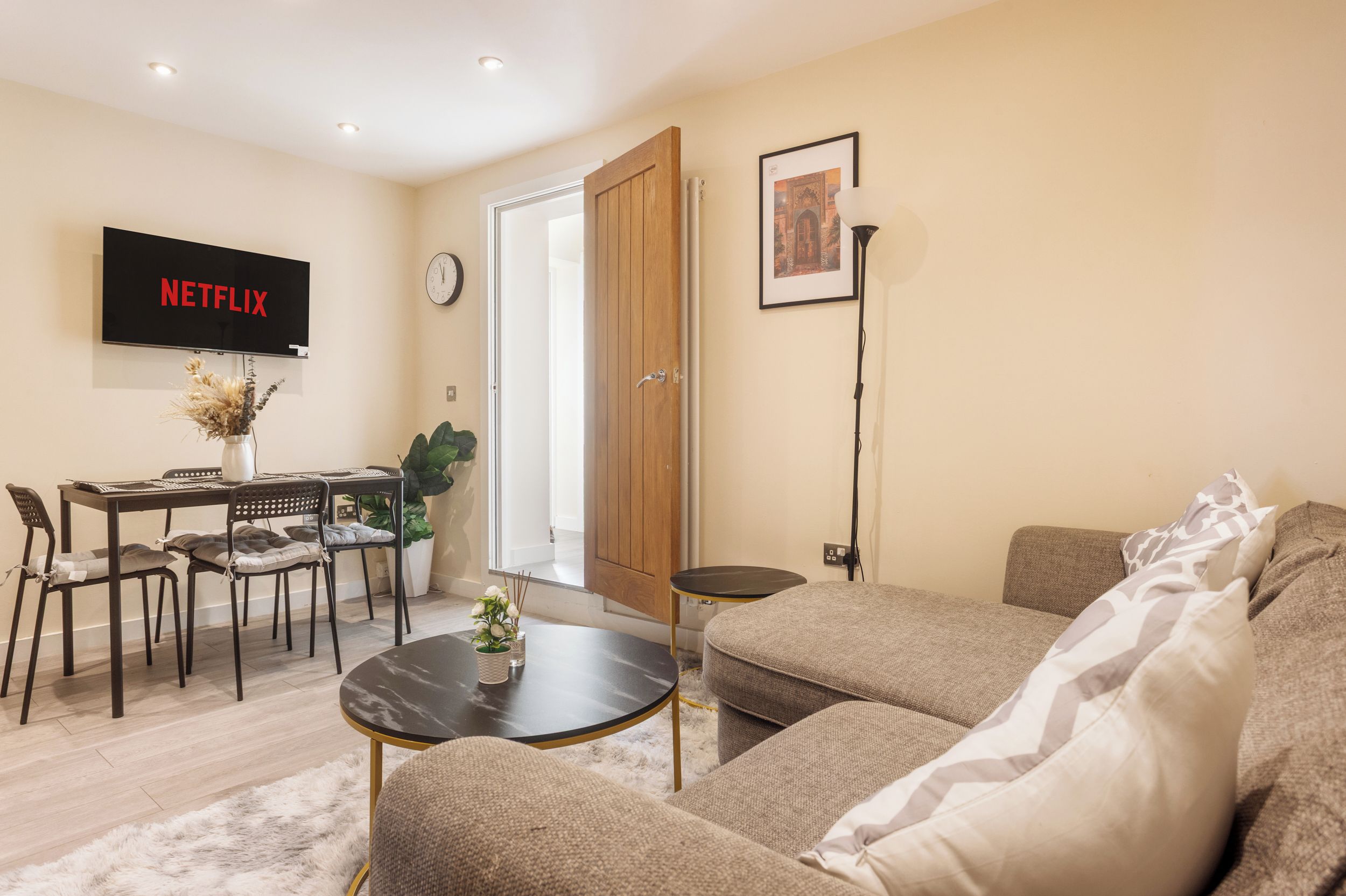 Een moderne woonkamer met een bank, een salontafel, een eettafel met stoelen, een aan de muur bevestigde televisie en een deur naar een andere kamer.