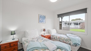 A bedroom with two beds, a nightstand, a lamp, a painting, and a window with a view of a building. - Phillip Island Cottage 6 (Cowes)