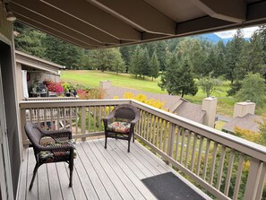 A wooden deck with two wicker chairs and a small table, overlooking a grassy area with trees and a distant mountain range. - Cozy 2 bedroom condo in amazing Mount Hood Village with AC on the golf course. (Mount Hood Village)
