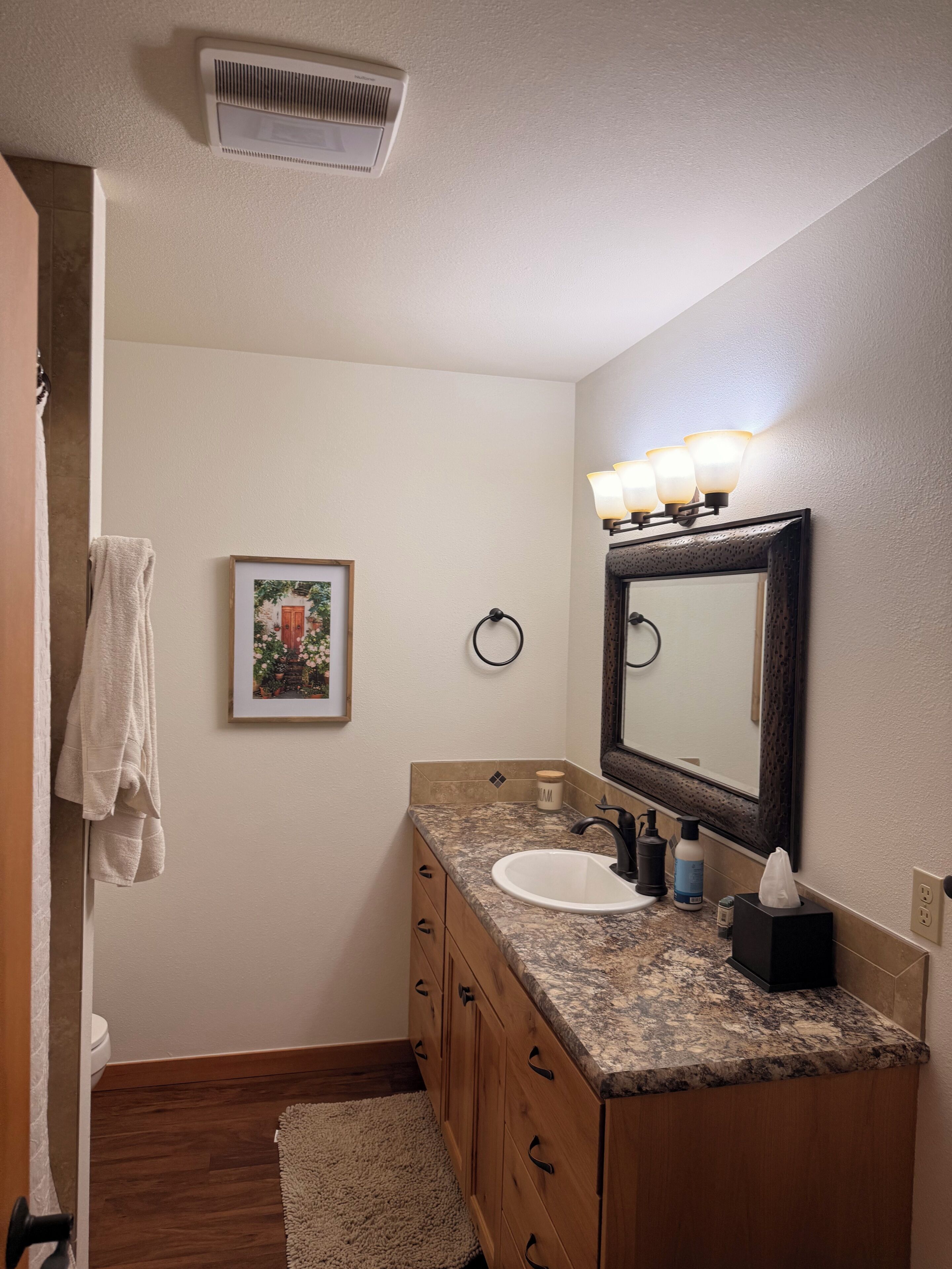 A bathroom with a white sink, a mirror with lights, a towel rack, and a framed picture on the wall.
