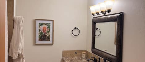 A bathroom with a white sink, a mirror with lights, a towel rack, and a framed picture on the wall.