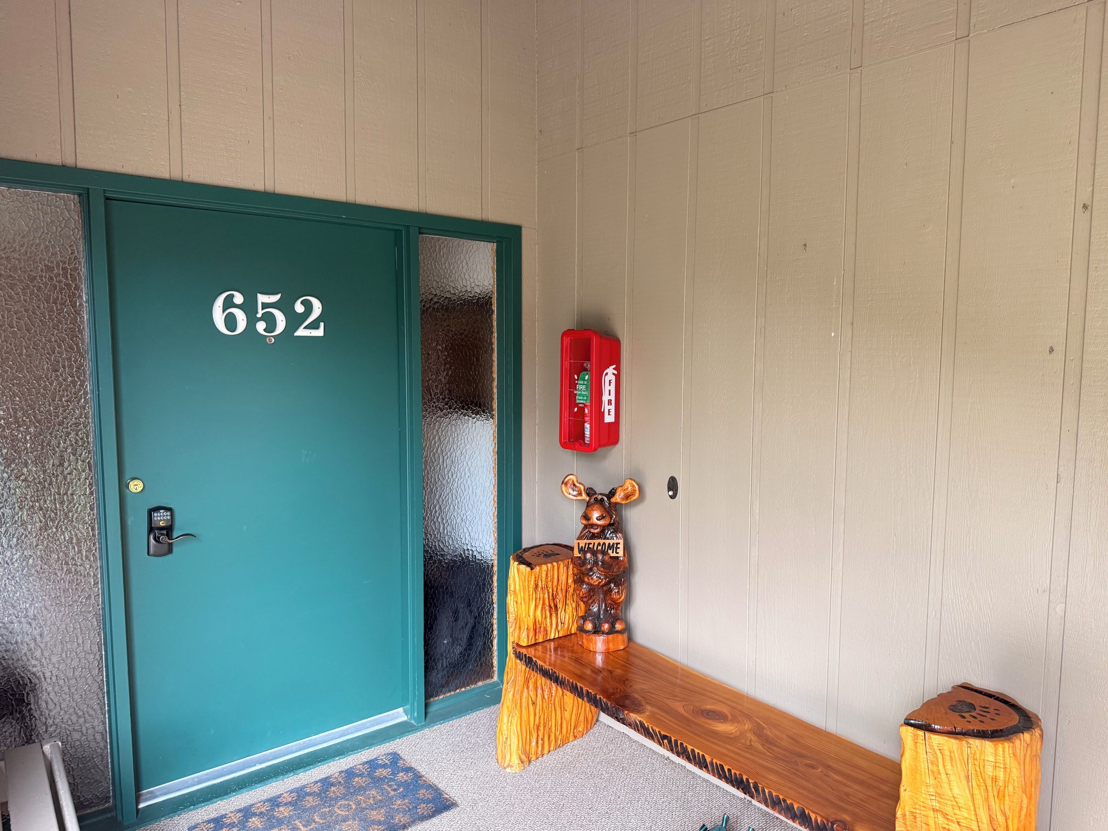 A teal door with the number 652, a fire alarm, and a wooden bench with a welcome sign.