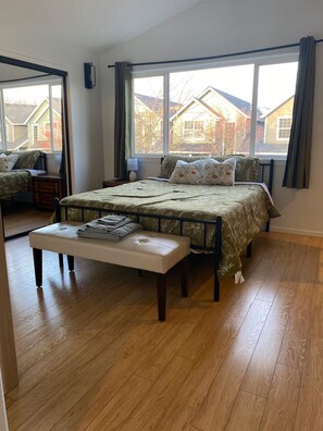 Een slaapkamer met een bed, een bankje, een raam en uitzicht op huizen.