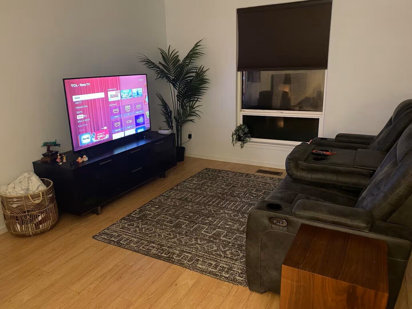 Una sala de estar con un televisor de pantalla plana sobre un pedestal negro, un tapete marrón estampado, un sofá gris y una ventana con persiana oscura.