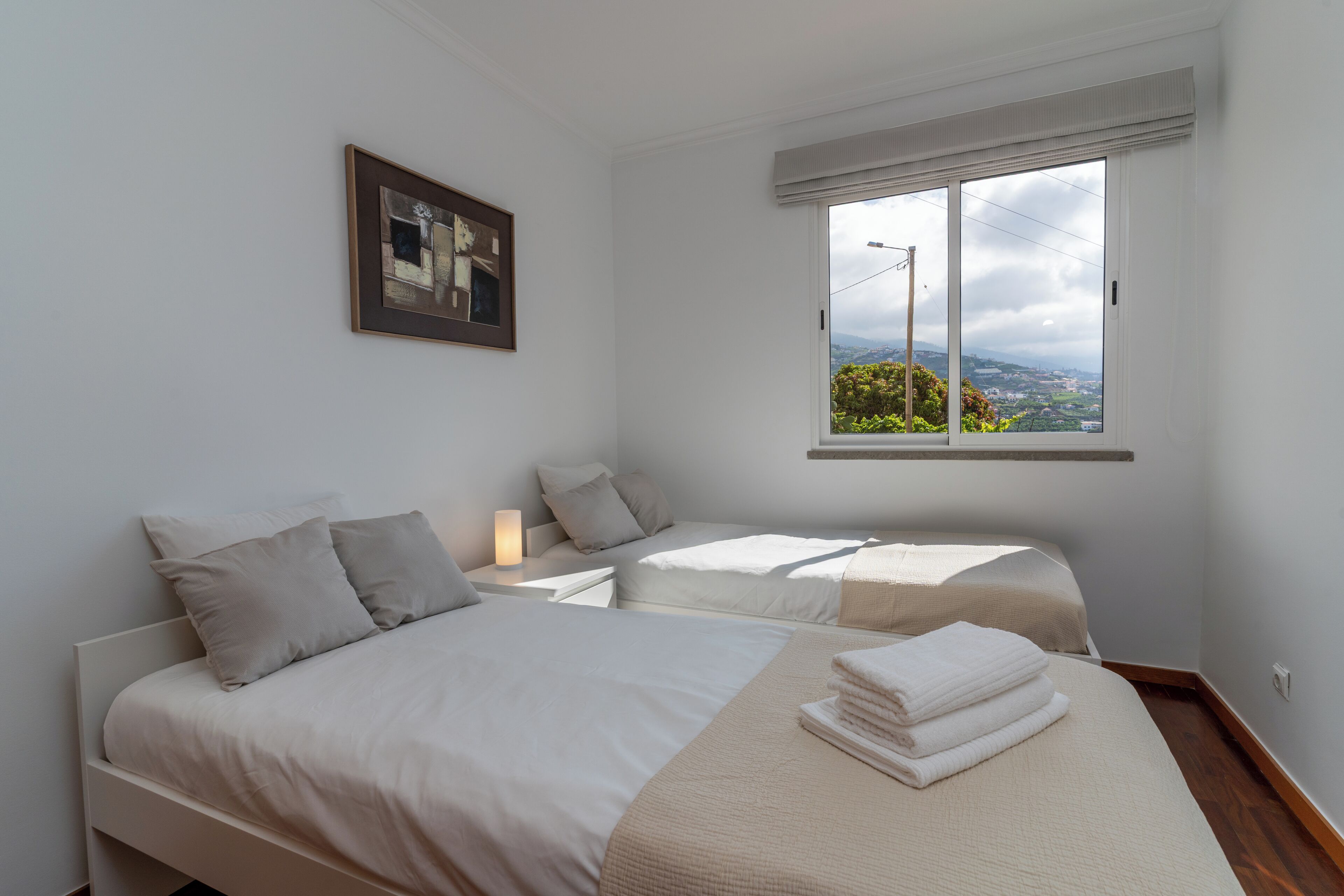 Ein Schlafzimmer mit zwei Betten, einem Nachttisch, einer Lampe, einem Fenster mit Blick und einem gerahmten Bild an der Wand.