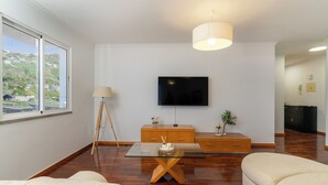 A modern living room with a flat-screen TV mounted on the wall, a wooden coffee table, a sofa, and a lamp. - Vrbo Property (Câmara de Lobos)