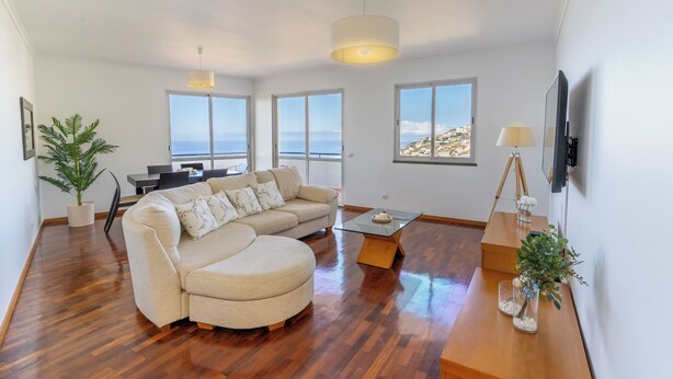 A spacious living area with a large sofa, a dining table, and a coffee table. There are multiple windows offering views of the sea and a hilly landscape. - Vrbo Property (Câmara de Lobos)