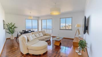 Ein großzügiger Wohnbereich mit einer großen Couch, einem Esstisch und einem Couchtisch. Mehrere Fenster bieten Ausblick auf das Meer und eine hügelige Landschaft.