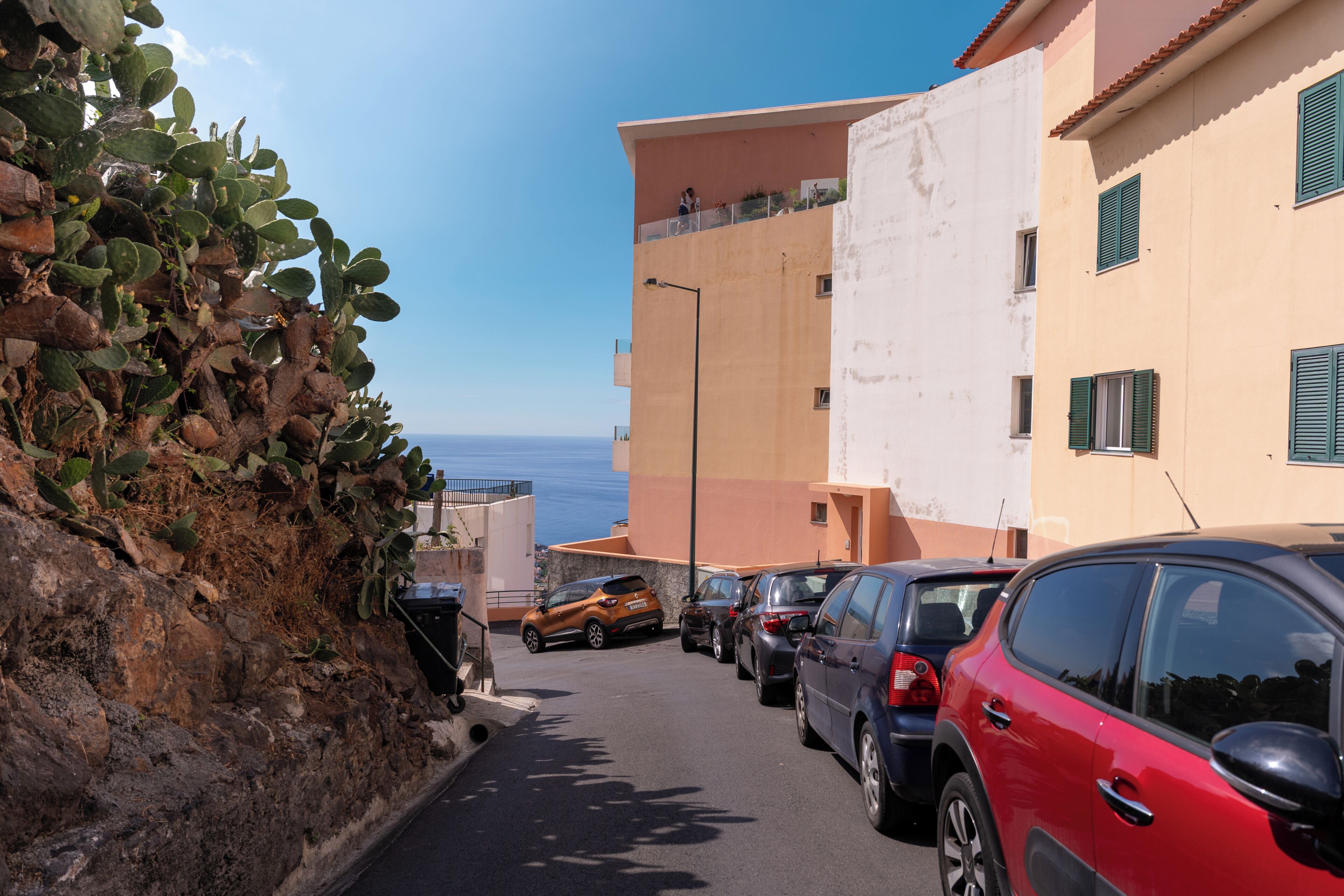 Eine schmale Straße mit parkenden Autos, Gebäuden mit Balkonen und freiem Blick auf das Meer.