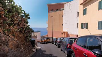 Eine schmale Straße mit parkenden Autos, Gebäuden mit Balkonen und freiem Blick auf das Meer.