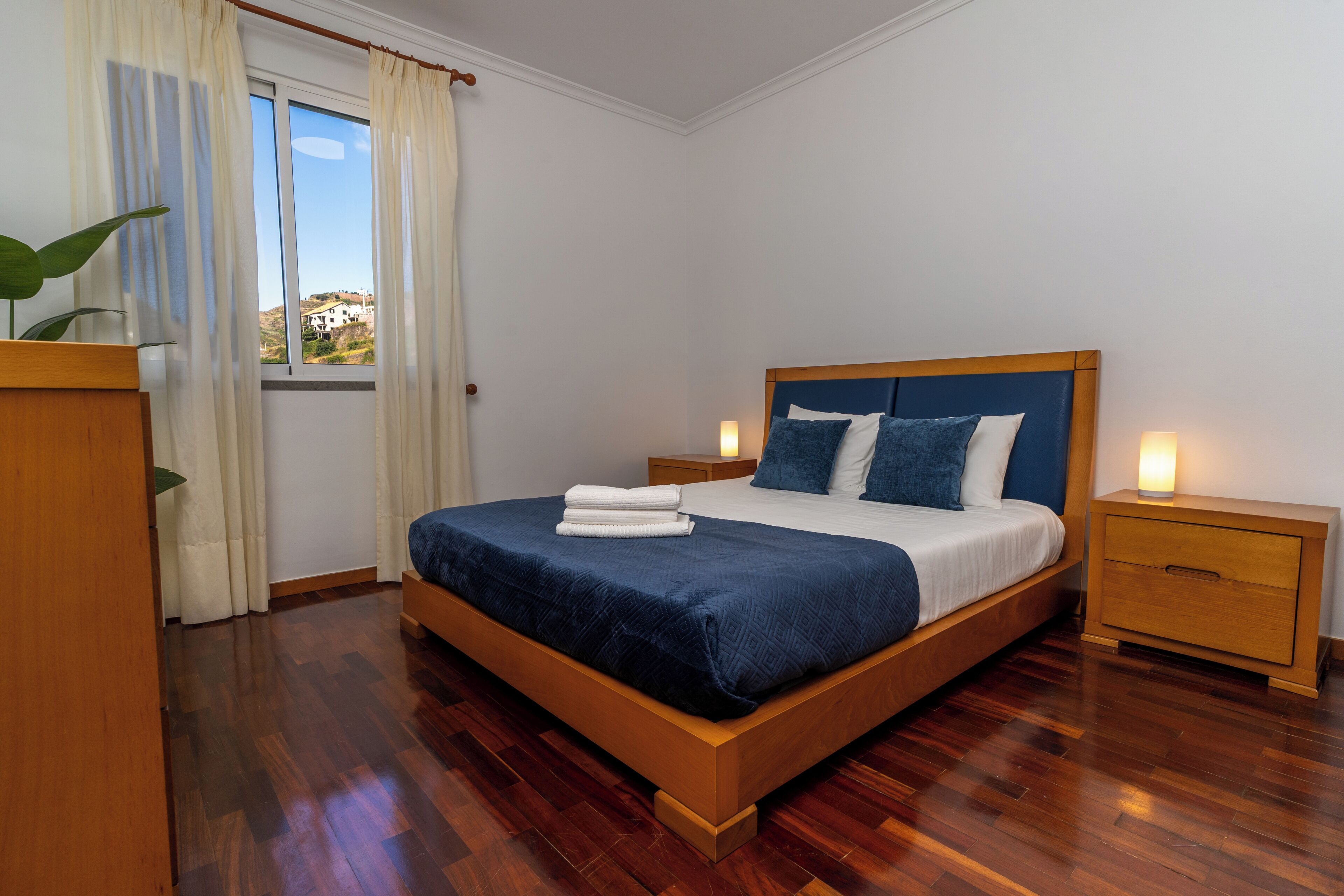 Ein ordentlich bezogenes Bett mit blauer Tagesdecke und weißen Leinen, flankiert von zwei Nachttischen mit Lampen, in einem Zimmer mit Holzboden und Blick durch ein Fenster.