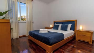 Ein ordentlich bezogenes Bett mit blauer Tagesdecke und weißen Leinen, flankiert von zwei Nachttischen mit Lampen, in einem Zimmer mit Holzboden und Blick durch ein Fenster.