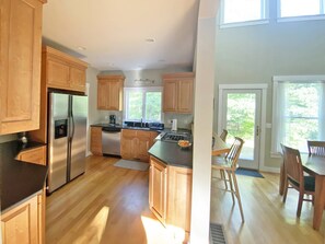 A modern kitchen with wooden cabinets, stainless steel appliances, and a dining area with wooden furniture. - Family Friendly Hidden Oaks Home (Oak Bluffs)