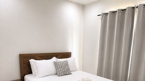 Un lit bien fait, recouvert d’une courtepointe blanche matelassée, avec une tête de lit en bois et deux oreillers. On y trouve une table de chevet en bois, dotée d’un tiroir, sur laquelle est posée une serviette enroulée. La pièce a des murs de couleur claire, un tapis à motifs foncés et une fenêtre avec des rideaux gris.