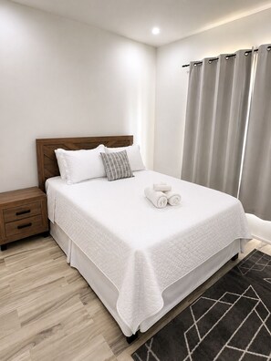 Ein ordentlich gemachtes Bett mit weißer, gesteppter Tagesdecke, einem Holz-Kopfteil und zwei Kissen. Ein Nachttisch aus Holz mit einer Schublade und einem gerollten Handtuch steht daneben. Der Raum hat helle Wände, einen dunkel gemusterten Teppich und ein Fenster mit grauen Vorhängen.