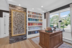 Una habitación con una estantería, un escritorio con un micrófono y una ventana con vista a árboles.