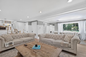 A spacious living room with two beige sofas, a wooden coffee table, and a view of a garden through a large window.