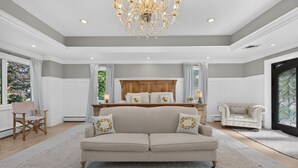 A spacious living room with a large beige sofa, a wooden headboard, and a chandelier. - Luxury Home w/ Secret Room & Royal Master Suite (Wyckoff)