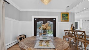 A dining room with a round wooden table, chairs, a vase with flowers, and a door with a decorative glass panel. - Luxury Home w/ Secret Room & Royal Master Suite (Wyckoff)