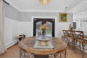 Uma sala de jantar com uma mesa de madeira redonda, cadeiras, um vaso com flores e uma porta com um painel de vidro decorativo.