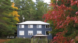 A two-story house with a visible attic window, surrounded by a well-maintained lawn and a large tree with red leaves. - The “Lower Dancehall”: A lakeview forested cottage (Port Sydney)