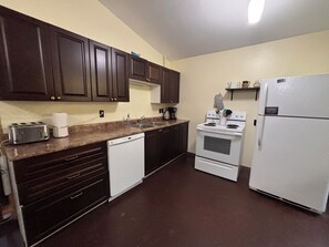 A kitchen with dark wooden cabinets, a white dishwasher, a stove, and a refrigerator. - The “Lower Dancehall”: A lakeview forested cottage (Port Sydney)