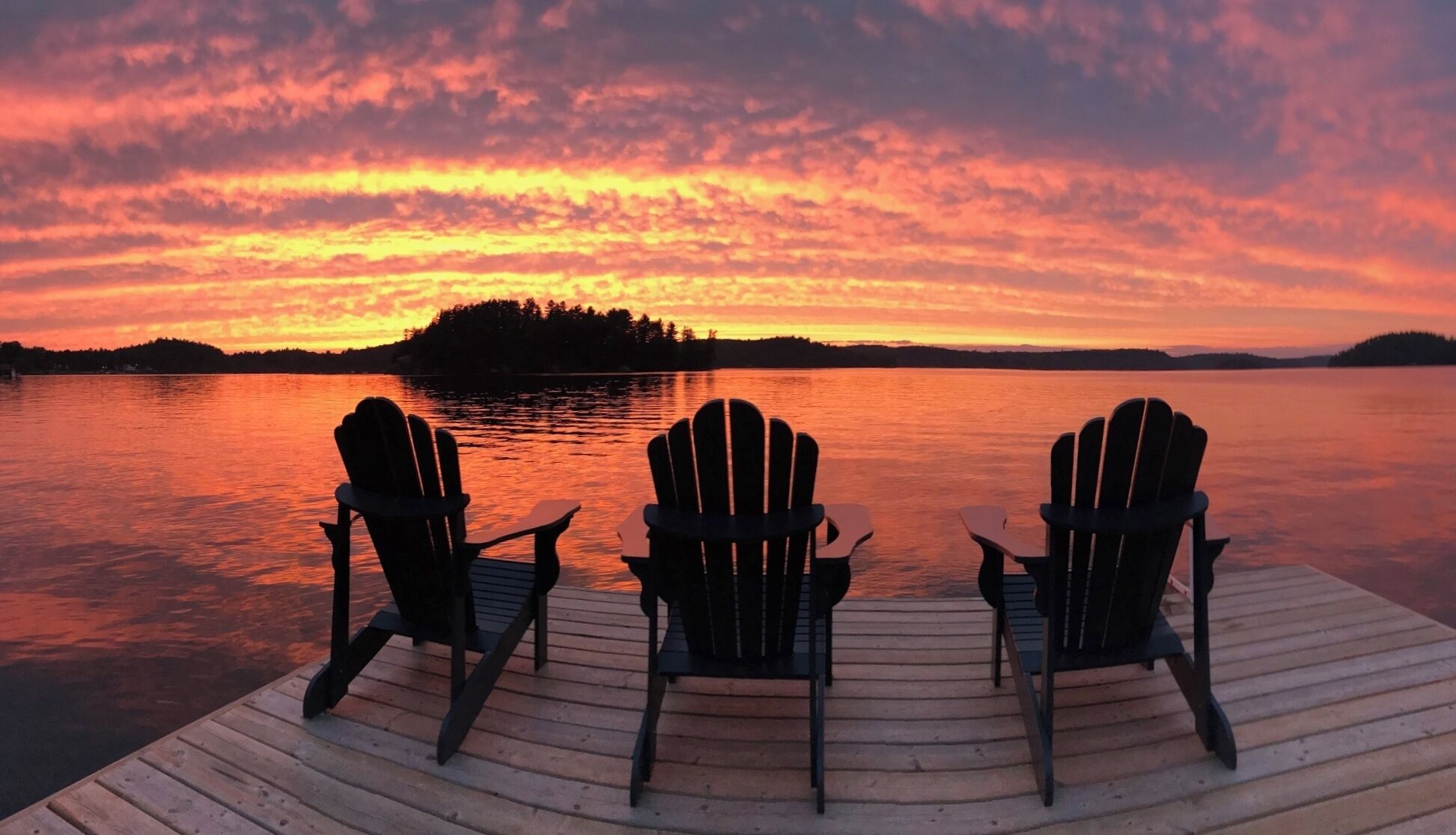 A wooden dock with three Adirondack chairs facing a sunset over a calm lake.