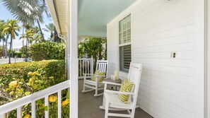 Eine Veranda mit weißen Geländern, zwei Schaukelstühle mit Kissen und Blick auf einen Garten mit Palmen und gelben Blüten.