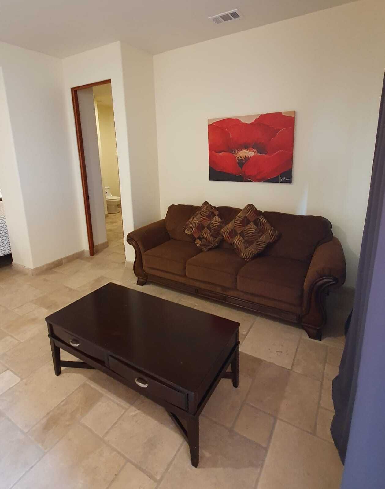 Uma sala de estar com um sofá marrom, uma mesa de centro de madeira escura e um quadro de uma flor vermelha na parede.