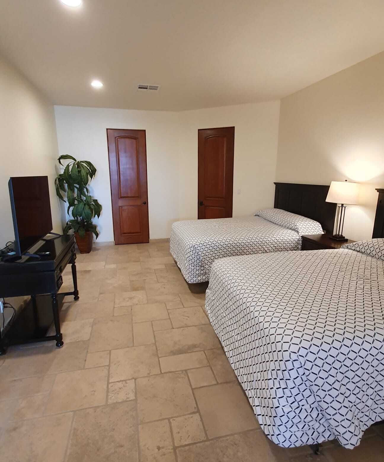 Quarto de hotel com duas camas, uma escrivaninha, uma luminária e um vaso de plantas.