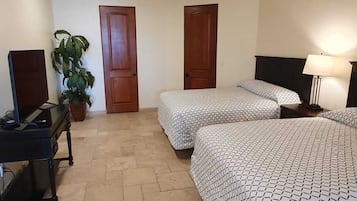 Quarto de hotel com duas camas, uma escrivaninha, uma luminária e um vaso de plantas.