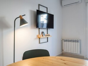 Une pièce comprenant une table ronde en bois, une chaise noire, un téléviseur fixé au mur, un lampadaire et un radiateur.