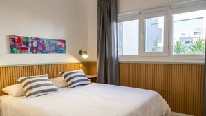 A hotel room with a bed, a window with curtains, a wall-mounted lamp, and a colorful abstract painting. - Apartment for 4 with a separate bedroom (Azul)