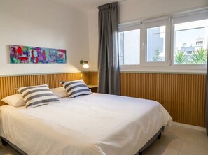 Une chambre d’hôtel avec un lit, une fenêtre avec des rideaux, une lampe fixée au mur et un tableau abstrait aux couleurs vives.