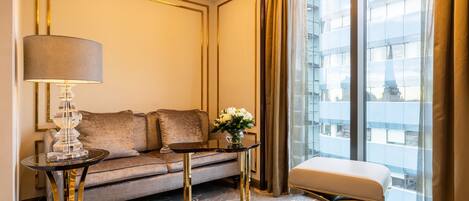 Habitació d'hotel moderna amb un sofà, una taula auxiliar, una làmpada i vista a un edifici per grans finestres.