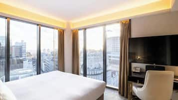 A hotel room with a large bed, a TV, and a city view through floor-to-ceiling windows.