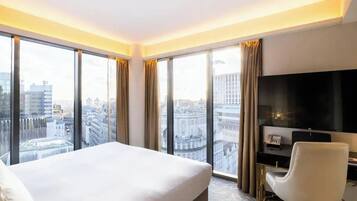 A hotel room with a large bed, a TV, and a city view through floor-to-ceiling windows.
