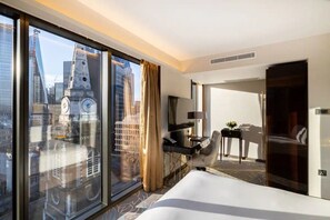 Een hotelkamer met een groot bed, een bureau met stoel, een televisie en uitzicht op een stadsgezicht door raam-tot-plafond ramen.
