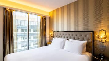 A hotel room with a large bed, a headboard with a tufted design, a nightstand with a clock, and a view of the city through the window.